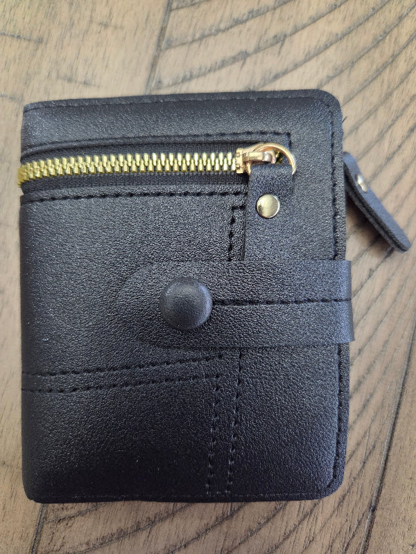 Black faux leather bifold wallet with a gold zipper, snap button closure, and a small gold rivet, shown on a wooden surface.