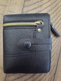 Black faux leather bifold wallet with a gold zipper, snap button closure, and a small gold rivet, shown on a wooden surface.