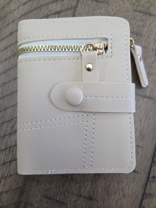 A small ivory colored faux leather zippered wallet with a snap strap closure and gold-tone zipper, shown on a dark wood background.