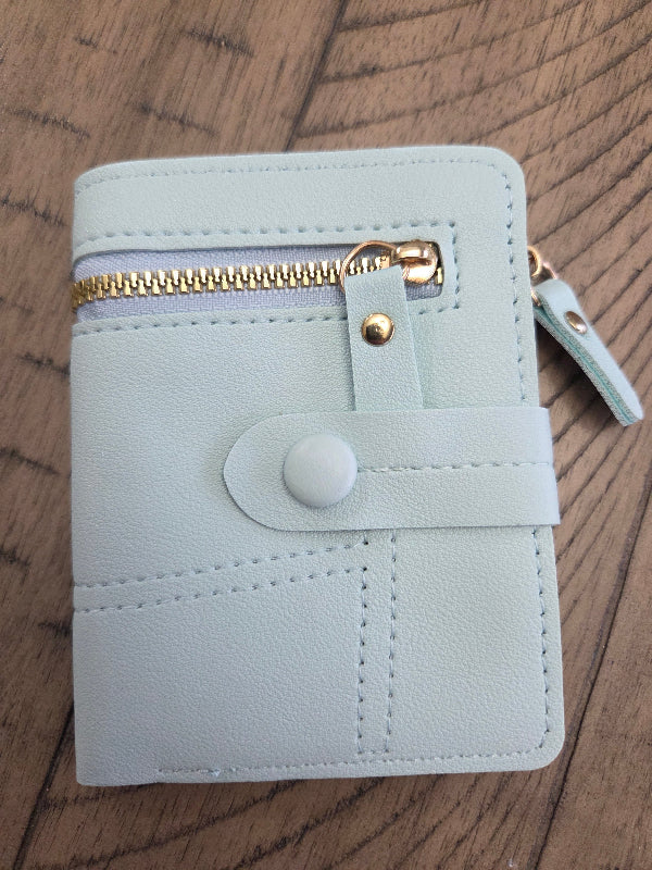 A light mint green faux leather wallet with a gold zipper, snap button closure, and a matching zip pull, shown on a dark wood background.