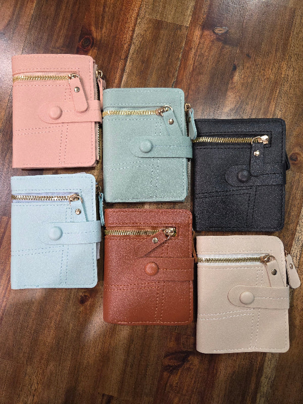 Six small zip-around wallet cases arranged on a wooden floor in assorted colors: pink, mint, black, light blue, rust brown, and cream. Each wallet has a snap button closure, a front card pocket, and a zip compartment.