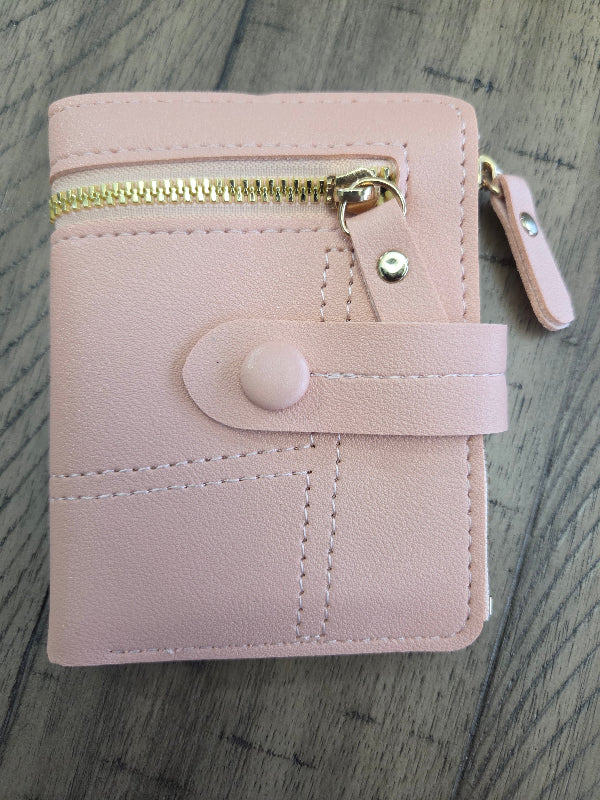 A light pink faux leather wallet with a gold zipper, snap button strap, and a small hanging tag, shown on a gray wood-textured background.