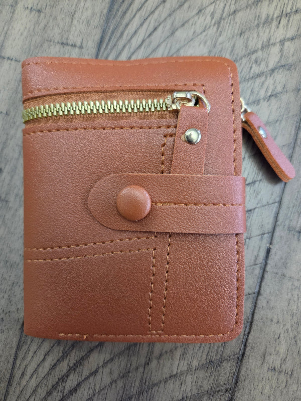 A rust orange colored faux-leather zip-around wallet with a snap-button strap closure and gold-tone metal hardware, photographed on a gray wood-grain surface.