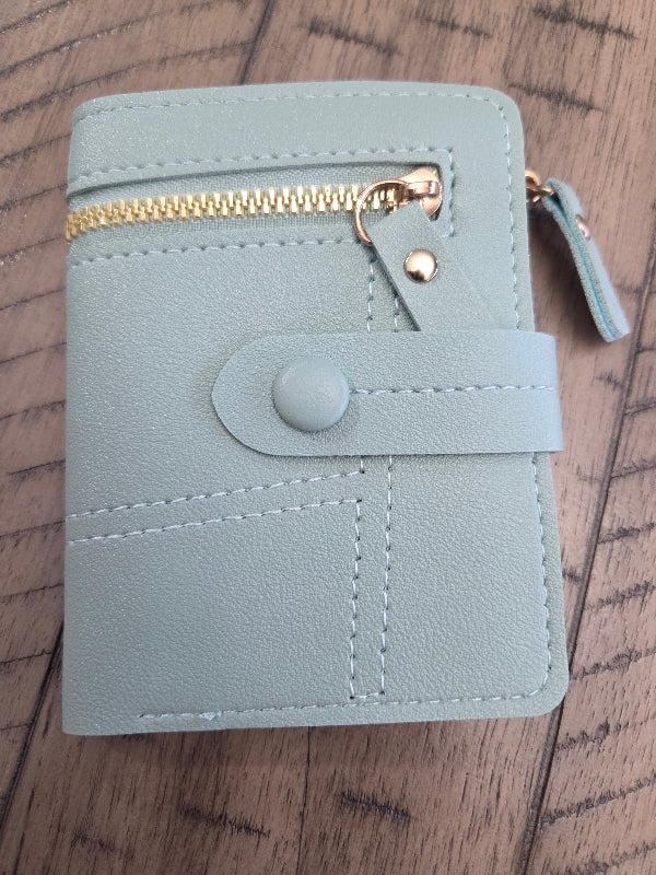 A light blue leather wallet with a gold zipper, snap button closure, and a small leather pull tab, shown on a wooden surface.